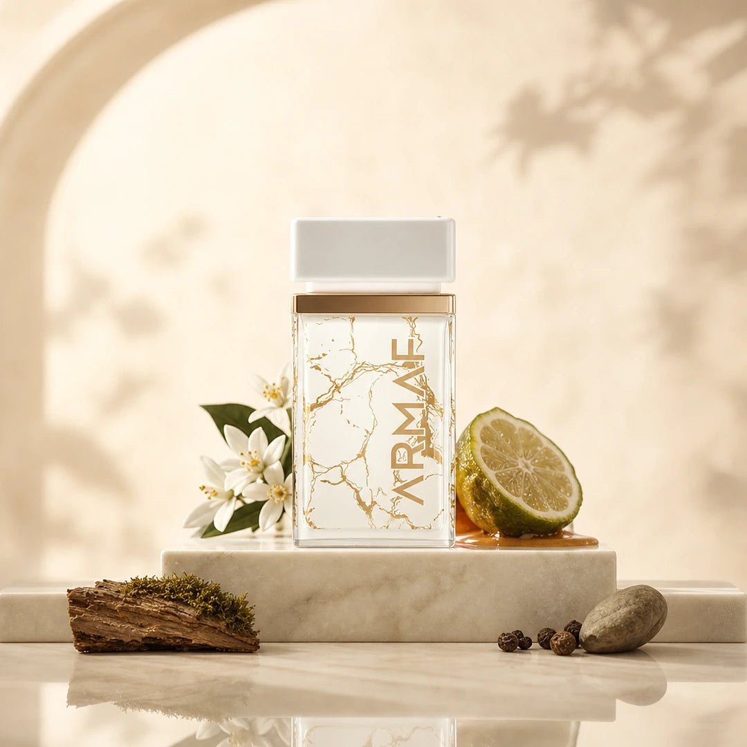 A rectangular clear perfume bottle with a white cap and gold marble-style veining, standing on a white marble pedestal. The bottle is styled with a sliced lime, white flowers, mossy wood, and small stones against a warm beige background with soft shadow overlays.